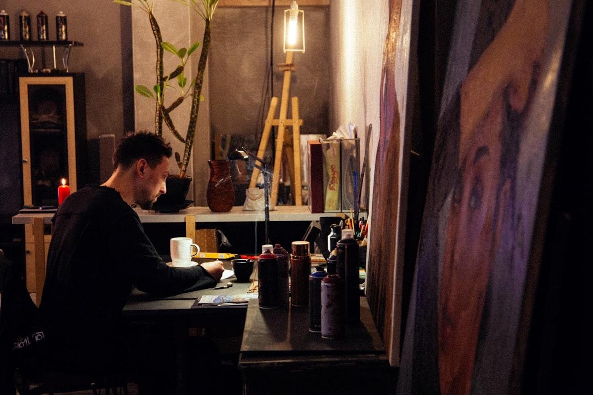 Artiste installé à sa table dans un atelier chaleureux, entouré de chevalets et de toiles peintes